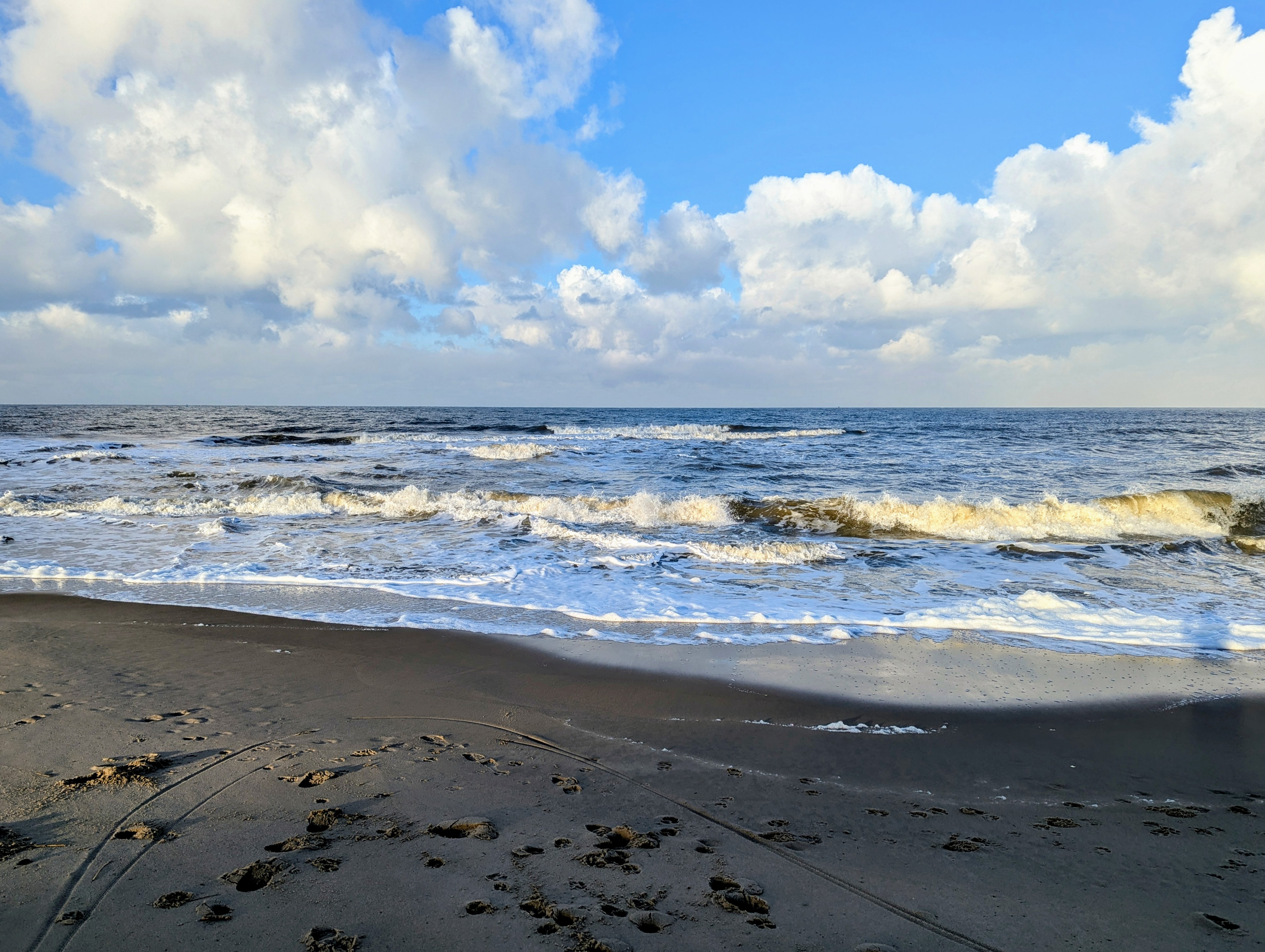 Sunlit waves rolling onto a sandy beach beneath an open blue sky and soft clouds
