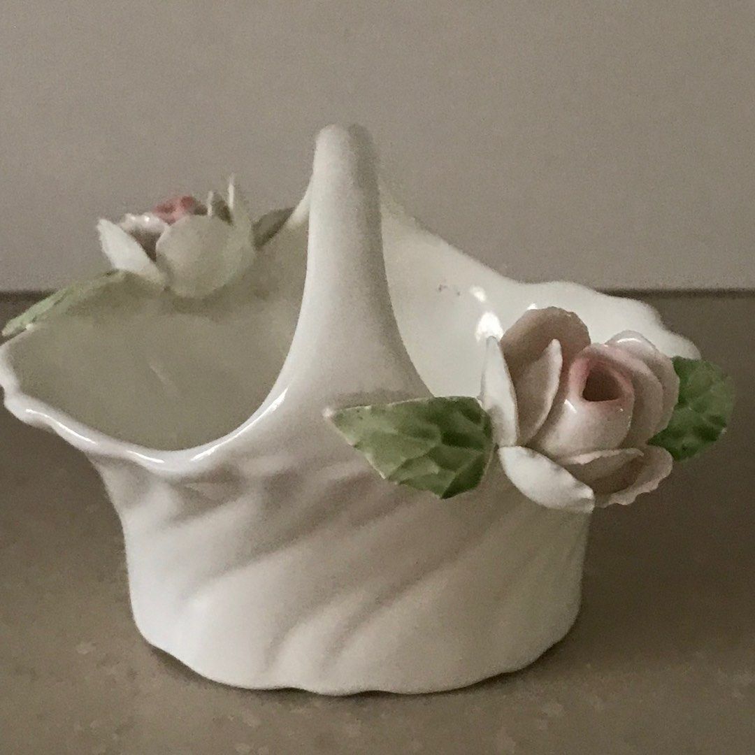 White ceramic basket with a tall arched handle, rippled sides, and sculpted pale pink roses with green leaves.