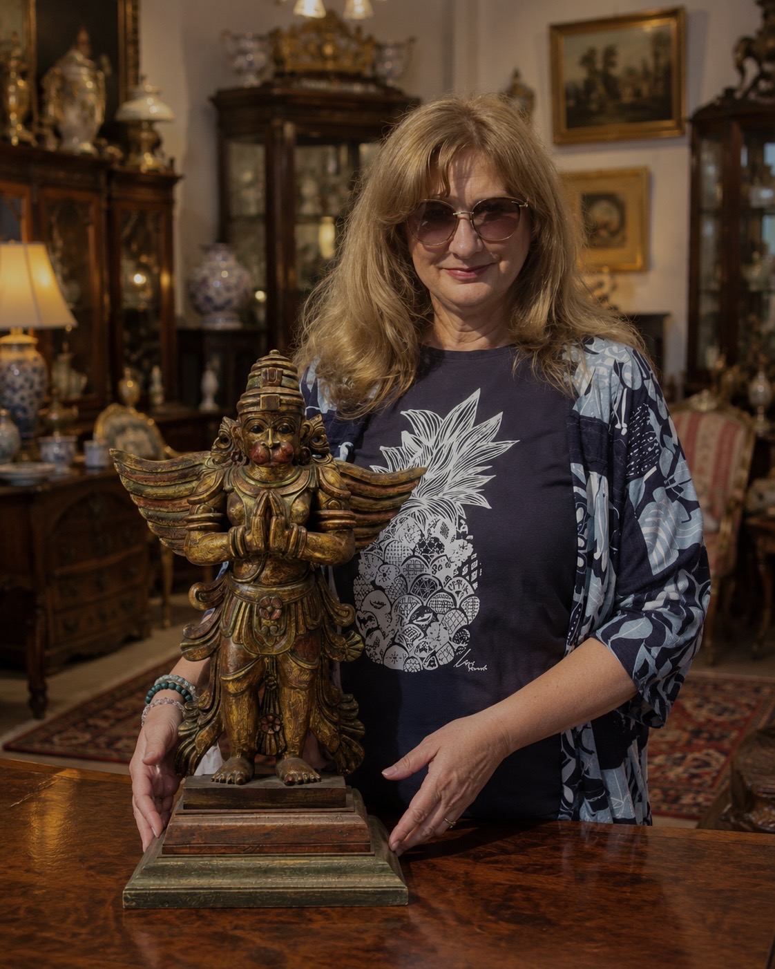 Woman presenting a gilded winged sculpture in an antique-filled room with wooden cabinets, porcelain, and framed paintings.