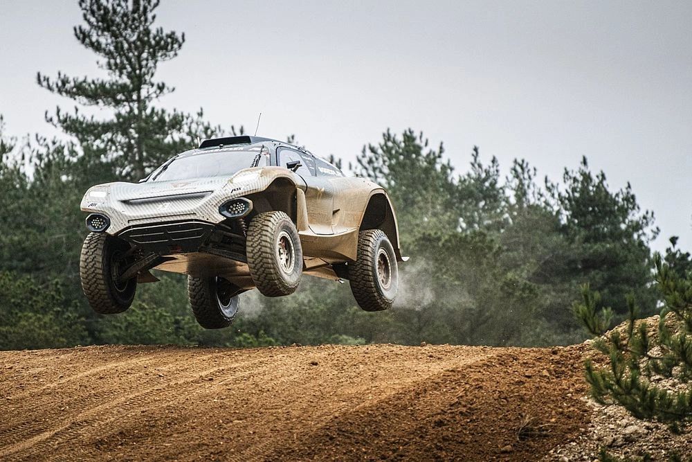 Electric off-road rally vehicle airborne above a dirt track, surrounded by pine trees.